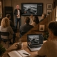 A PixelSchool 2026/27 - Hybrid - Vor-Ort und Online Kursleiter unterrichtet eine kleine Gruppe im gemütlichen Raum mit Brückenfoto auf Großbildschirm. Inset: derselbe Hybrid-Unterricht läuft online auf einem Laptop neben Kaffee und Notizbuch am Holztisch.