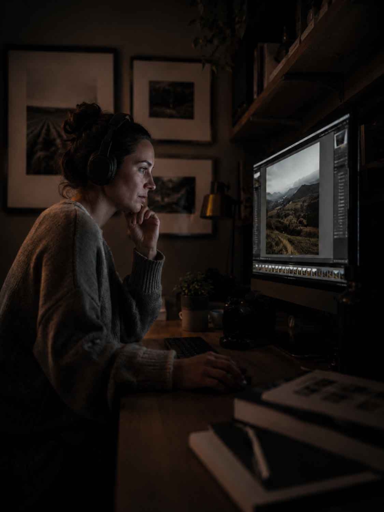 Eine Frau mit Kopfhörern sitzt an einem Schreibtisch in einem schwach beleuchteten Raum und bearbeitet ein Landschaftsfoto an ihrem Computer - eine ideale Umgebung, um in einen Bildbearbeitung Online-Kurs einzutauchen. Im Hintergrund sind Regale, gerahmte Bilder und Bücher zu sehen.