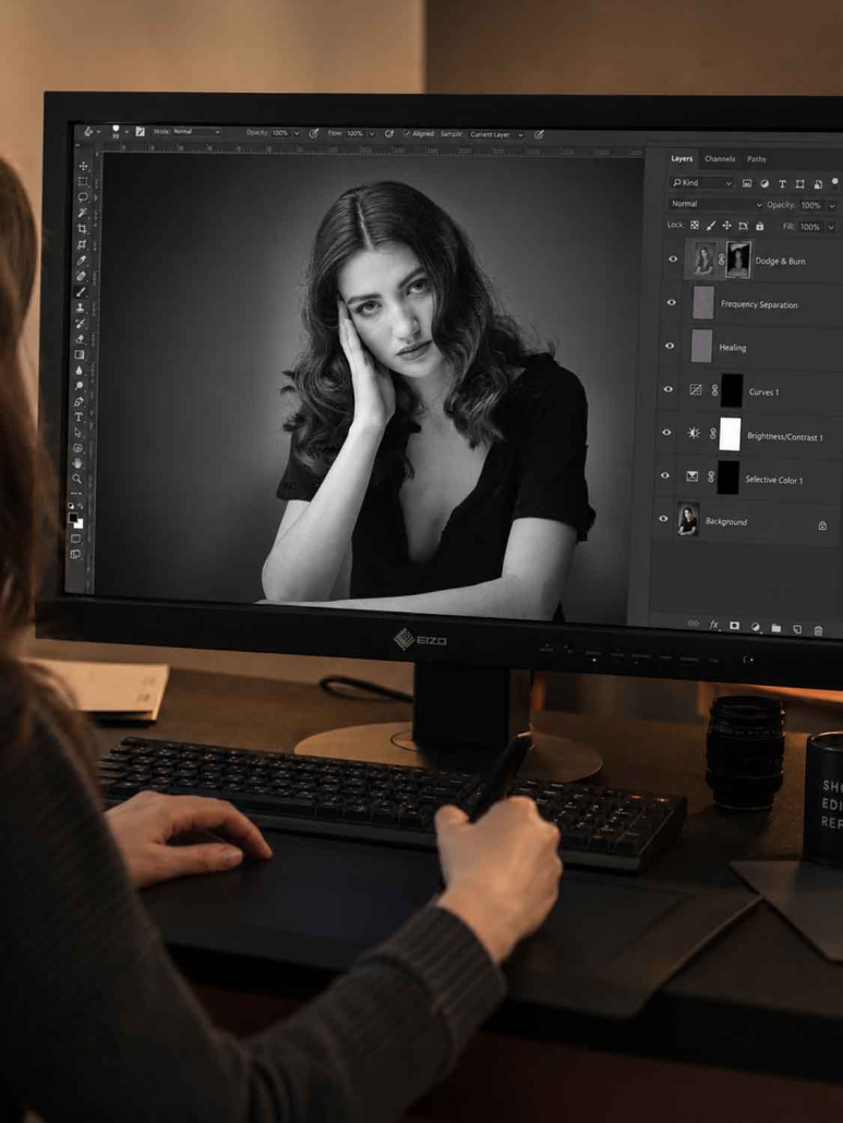 Eine Person bearbeitet mit einem Stift das Schwarz-Weiß-Porträt einer Frau auf einem Computermonitor, wobei Bearbeitungssoftware und Einstellungsebenen auf dem Bildschirm sichtbar sind.