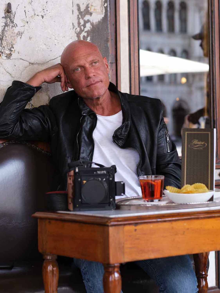 Frank Werner Fotograf, ein glatzköpfiger Mann in einer schwarzen Lederjacke, sitzt mit seiner Kamera, einem Glas Tee und einem Teller Chips an einem Cafétisch. Er stützt den Kopf auf seine Hand und blickt nachdenklich in die Ferne.