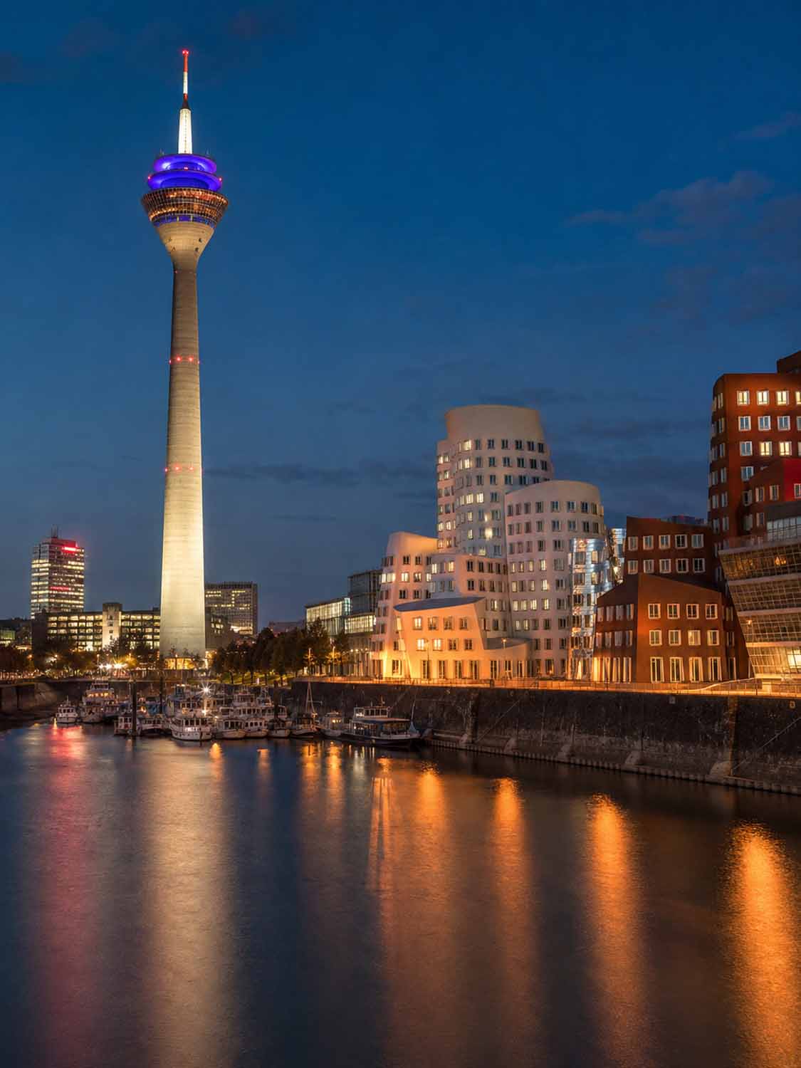 Nachtansicht des beleuchteten Düsseldorfer Rheinturms und des MedienHafens mit schimmernden Reflexionen - die perfekte Inspiration für einen Bildbearbeitung Workshop Düsseldorf entlang der pulsierenden Uferpromenade.