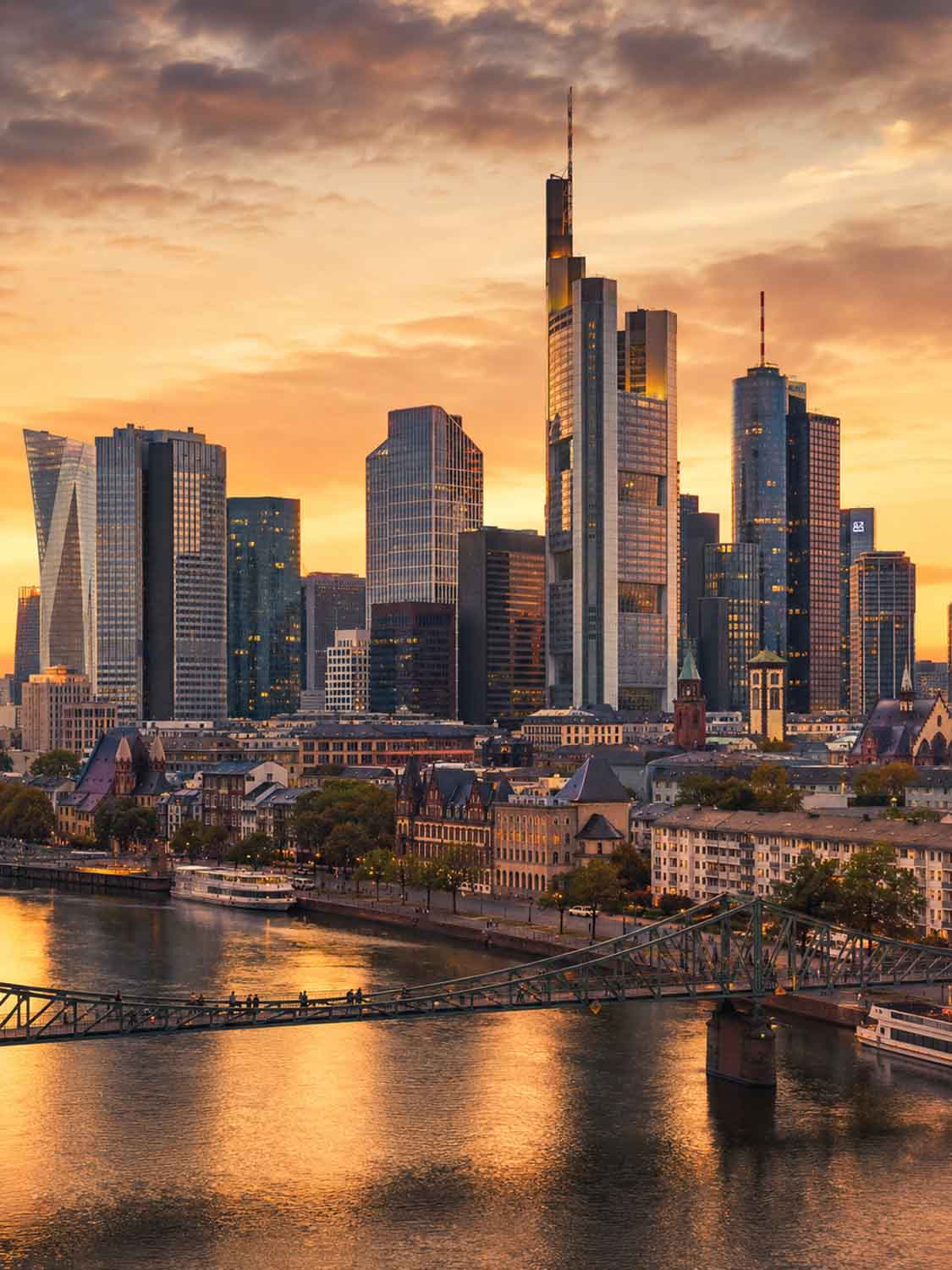 Eine Stadtsilhouette mit modernen Hochhäusern bei Sonnenuntergang, die sich in einem Fluss spiegelt. Der Himmel ist teilweise bewölkt, und eine Brücke führt über das Wasser - eine inspirierende Szene für jeden Bildbearbeitung Workshop Frankfurt.