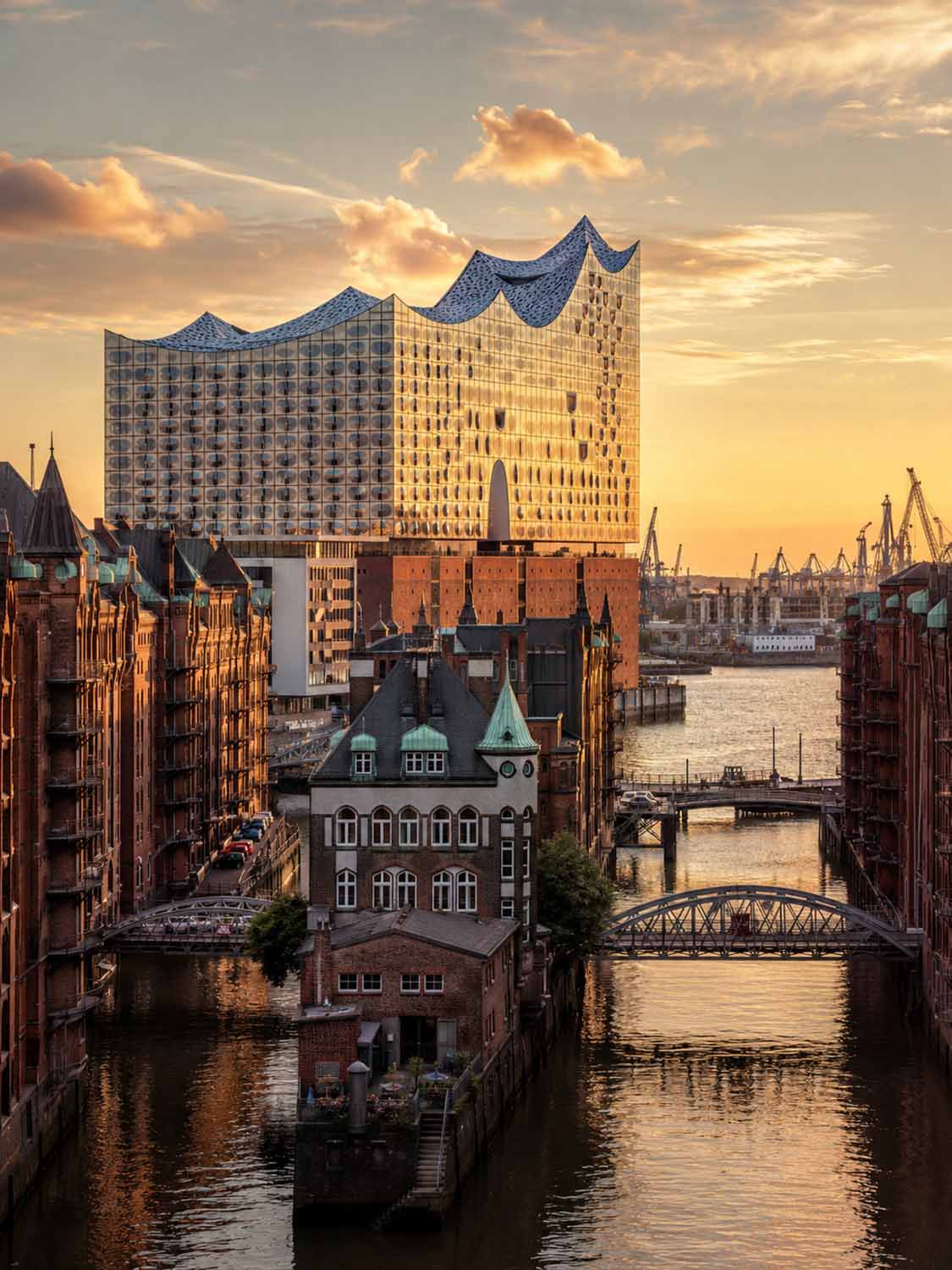 Ein Blick auf die Elbphilharmonie in Hamburg bei Sonnenuntergang, umgeben von historischen Backsteingebäuden, einem Kanal und Brücken, die goldenes Licht reflektieren - die perfekte Inspiration für einen Bildbearbeitung Workshop Hamburg.