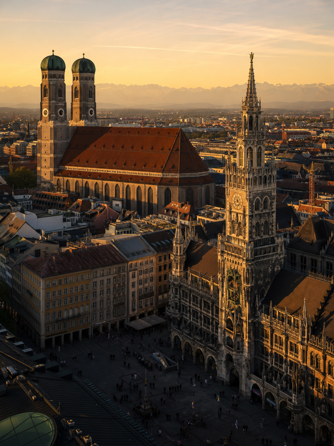 Luftaufnahme der Münchner Innenstadt bei Sonnenuntergang, mit der Frauenkirche mit ihren Zwillingskuppeln und dem Neuen Rathaus mit seinem kunstvollen Uhrenturm; die perfekte Inspiration für einen Bildbearbeitung Workshop München, mit entfernten Bergen unter einem goldenen Himmel.