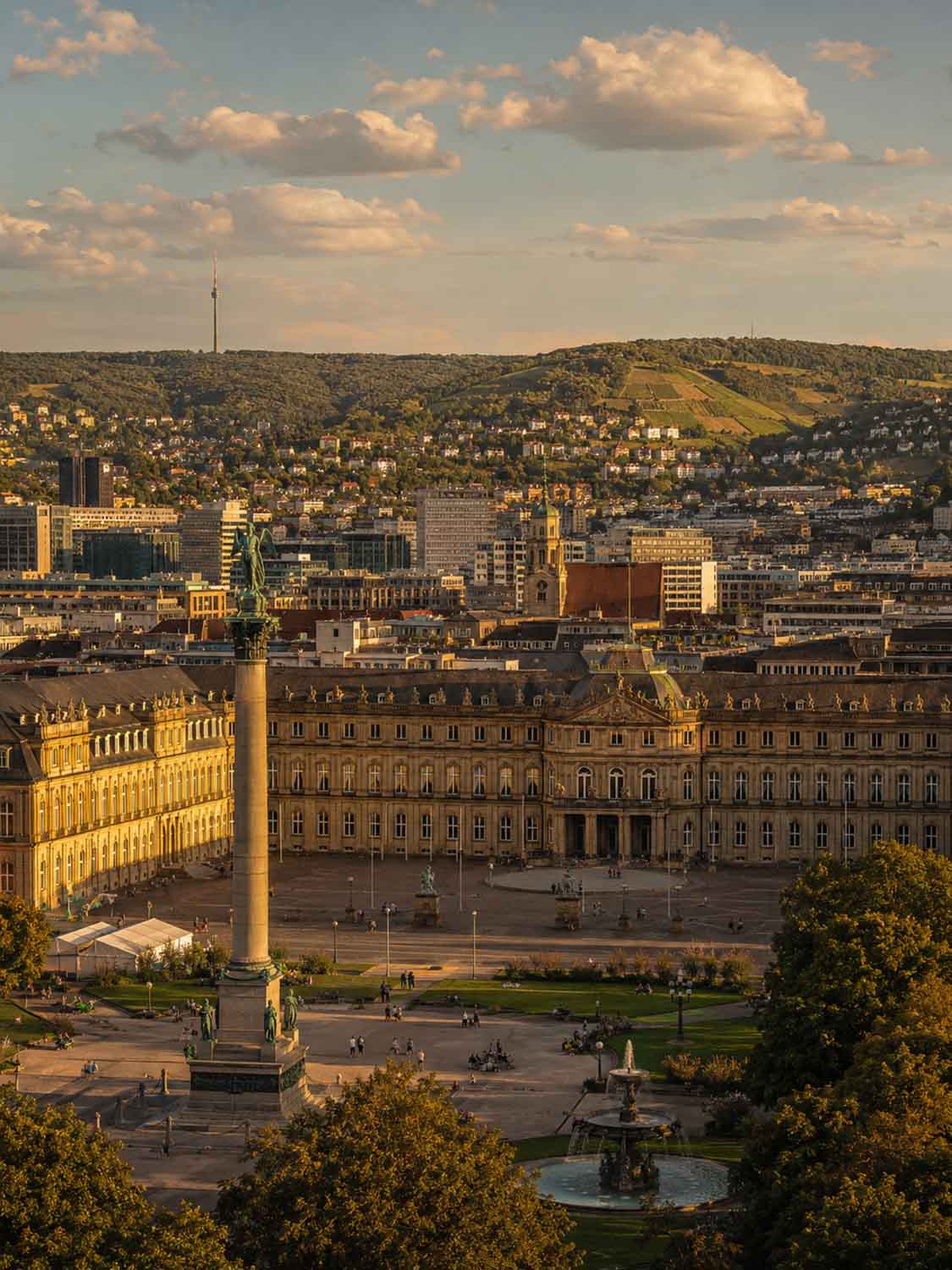 Luftaufnahme des Stuttgarter Schlossplatzes mit der Jubiläumssäule, dem Brunnen und den historischen Gebäuden - eine inspirierende Kulisse für einen Bildbearbeitung Workshop Stuttgart, eingebettet in grüne Hügel unter einem teilweise bewölkten Himmel.