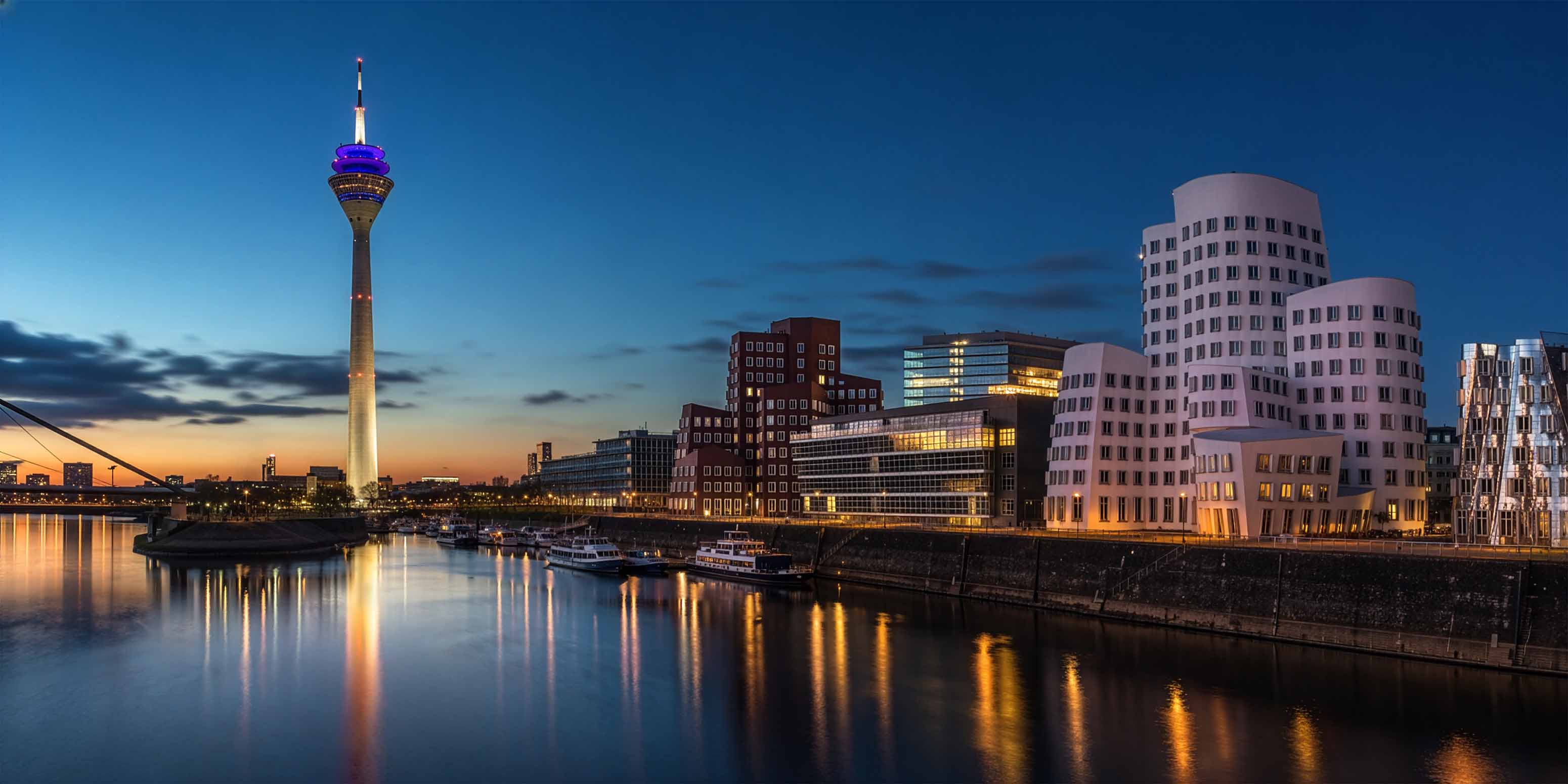 Nächtliche Ansicht der Düsseldorfer Skyline mit dem beleuchteten Rheinturm und den modernen Gebäuden am Wasser - eine inspirierende Szene für jeden Bildbearbeitung Workshop Düsseldorf, mit schimmernden Spiegelungen auf dem ruhigen Fluss unter dem Dämmerungshimmel.