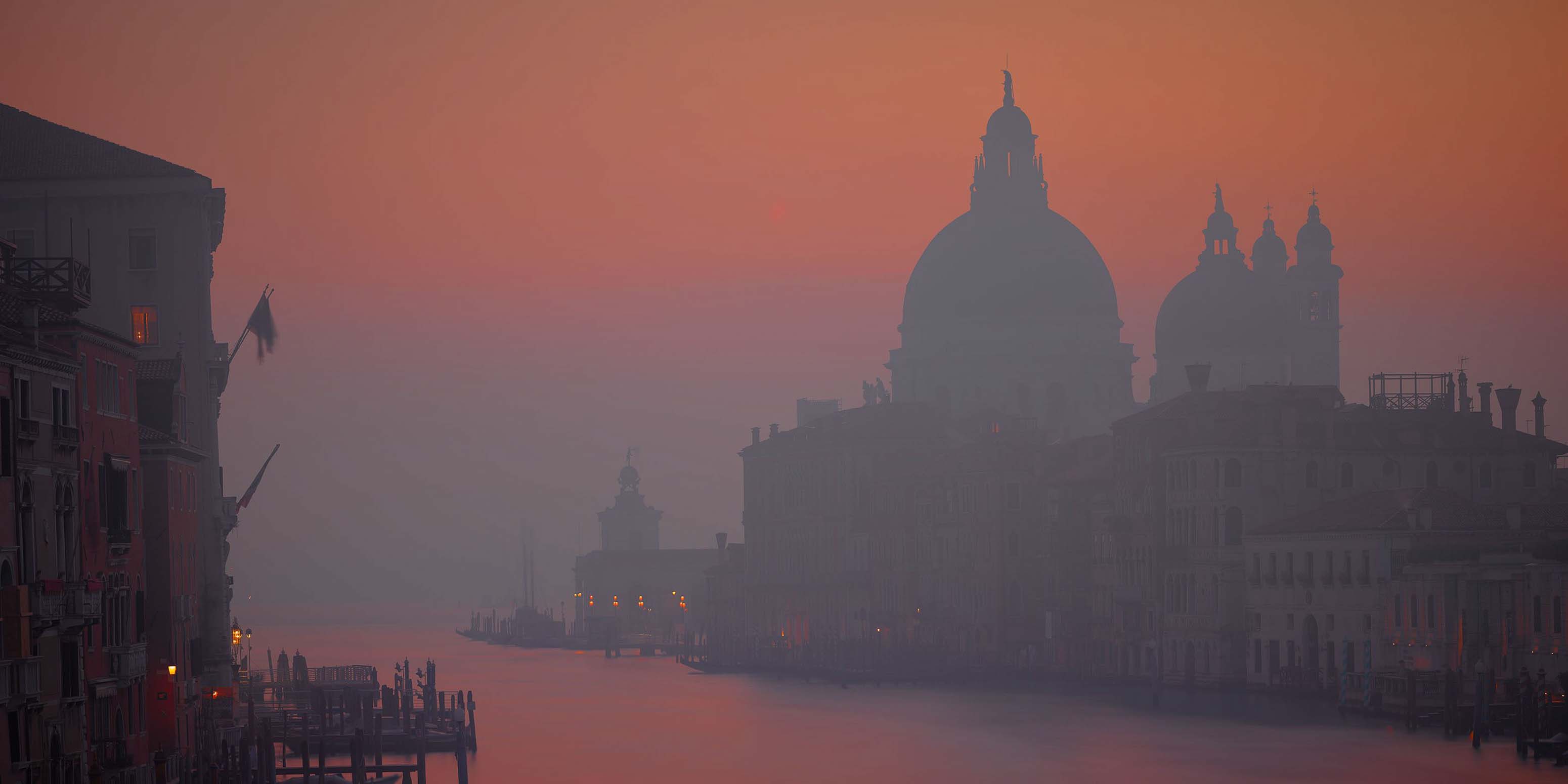 Ein dunstiger Sonnenuntergang taucht Venedig in ein orangefarbenes Licht, während die Silhouetten historischer Gebäude und Kuppeln einen ruhigen Kanal säumen.