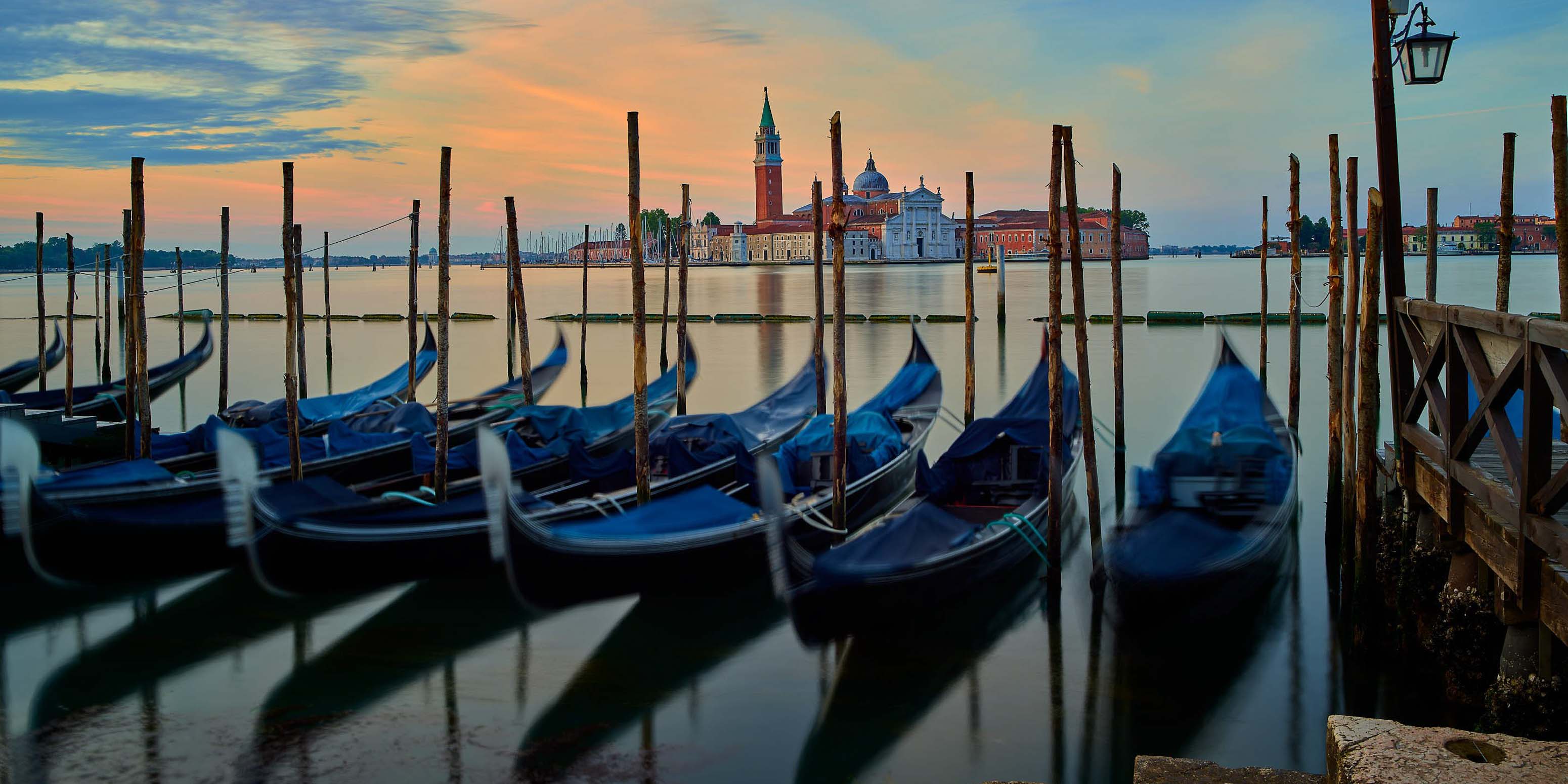 Blaue Gondeln, die an Holzpfählen festgemacht sind, schaukeln sanft auf einem ruhigen Kanal bei Sonnenuntergang, während auf der anderen Seite des Wassers in Venedig, Italien, die historische Insel San Giorgio Maggiore mit ihrer Kirche und ihrem Glockenturm zu sehen ist.
