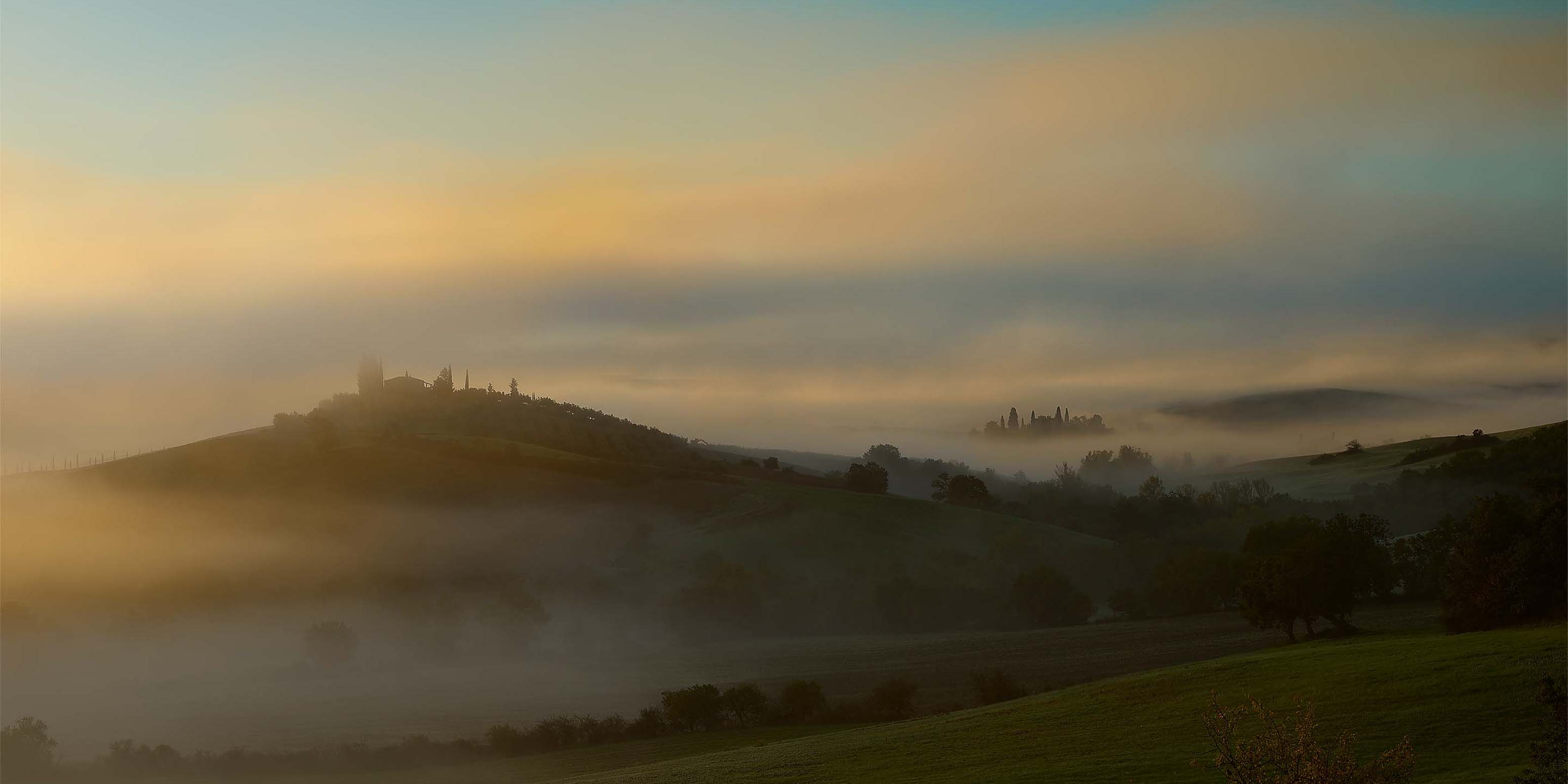 Ein nebliger Sonnenaufgang über sanften Hügeln, wobei die Silhouetten von Gebäuden und Bäumen durch den Nebel kaum zu erkennen sind und in sanftes goldenes und blaues Licht getaucht sind.