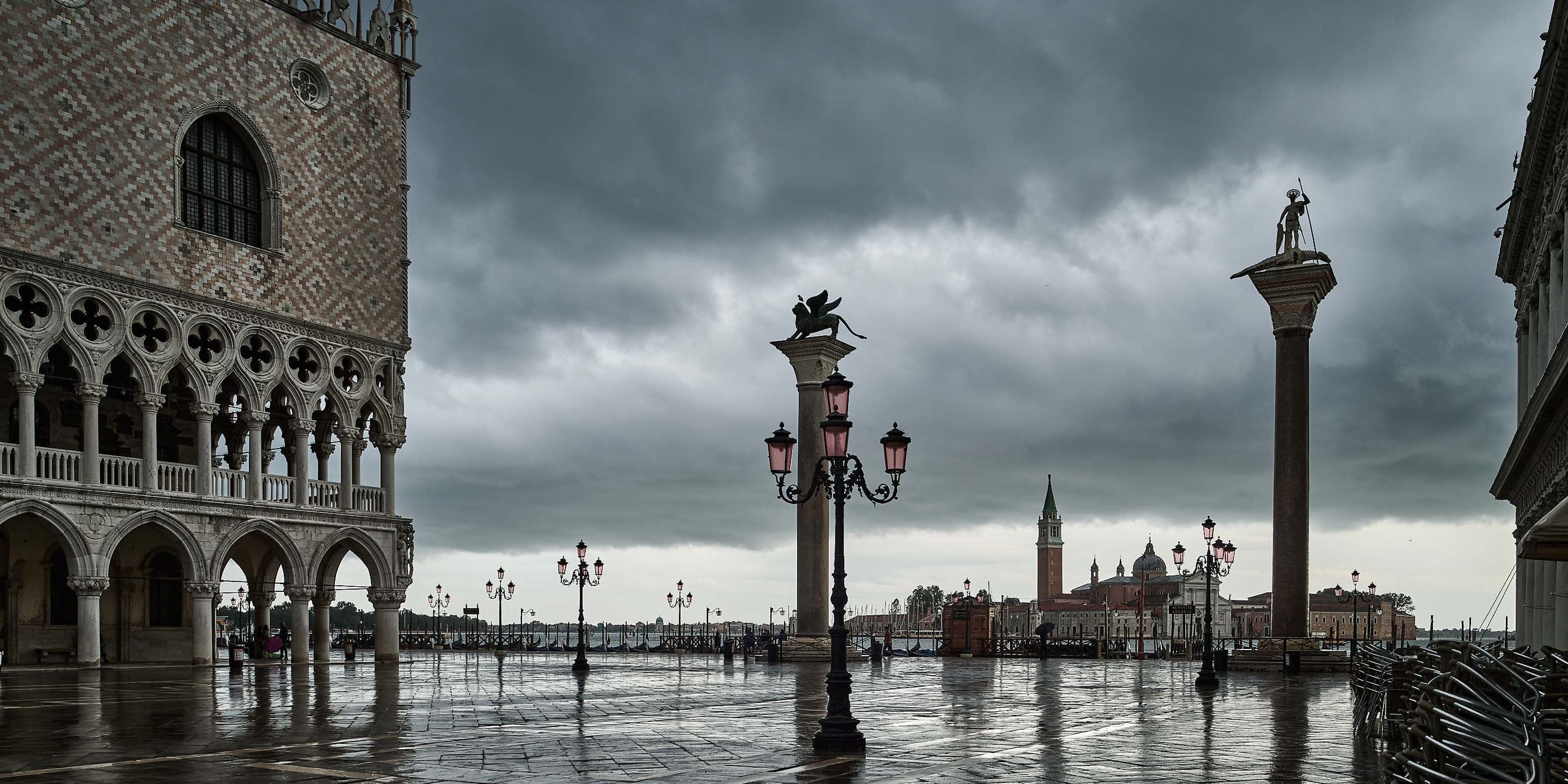 Ein dramatischer, bewölkter Himmel ragt über der Piazza San Marco in Venedig empor, mit nassen Kopfsteinpflastersteinen, kunstvollen Straßenlaternen, dem Dogenpalast auf der linken Seite und entfernten Türmen und Statuen, die sich über dem Wasser als Silhouetten abzeichnen.
