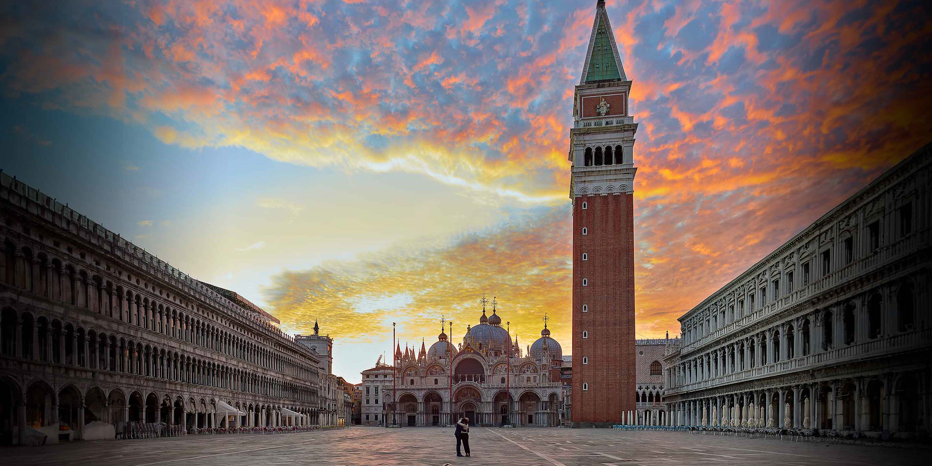 Ein Weitwinkelblick auf den Markusplatz in Venedig bei Sonnenaufgang, mit dem hohen Campanile, der Markusbasilika und den umliegenden historischen Gebäuden vor einem dramatischen, farbenprächtigen Himmel.