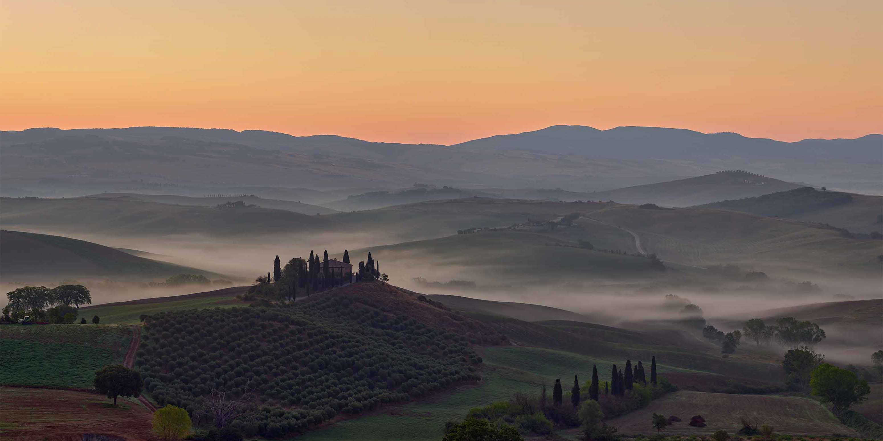 Sanfte Hügel mit vereinzelten Bäumen und Feldern liegen im Morgennebel unter einem sanften orange-rosa Sonnenaufgangshimmel, im Hintergrund sind ferne Berge zu sehen.