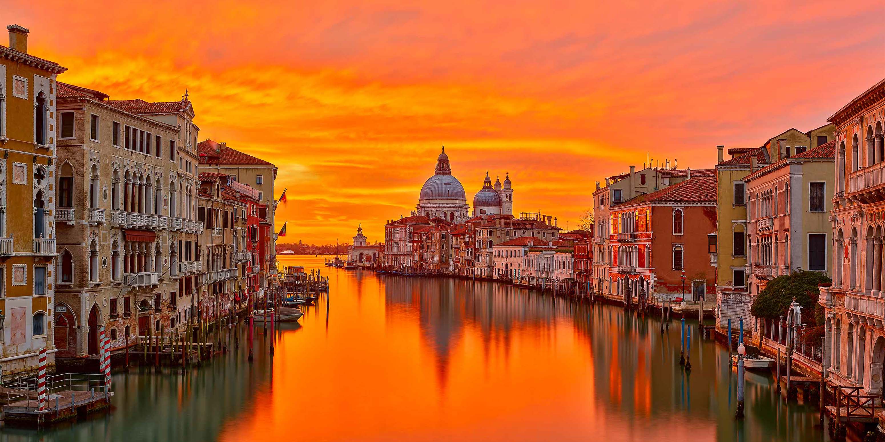 Ein leuchtend orangefarbener Sonnenuntergang erhellt den Himmel über dem Canal Grande in Venedig und spiegelt sich im Wasser wider, während auf beiden Seiten historische Gebäude stehen und im Hintergrund die Kuppel der Basilica di Santa Maria della Salute zu sehen ist.