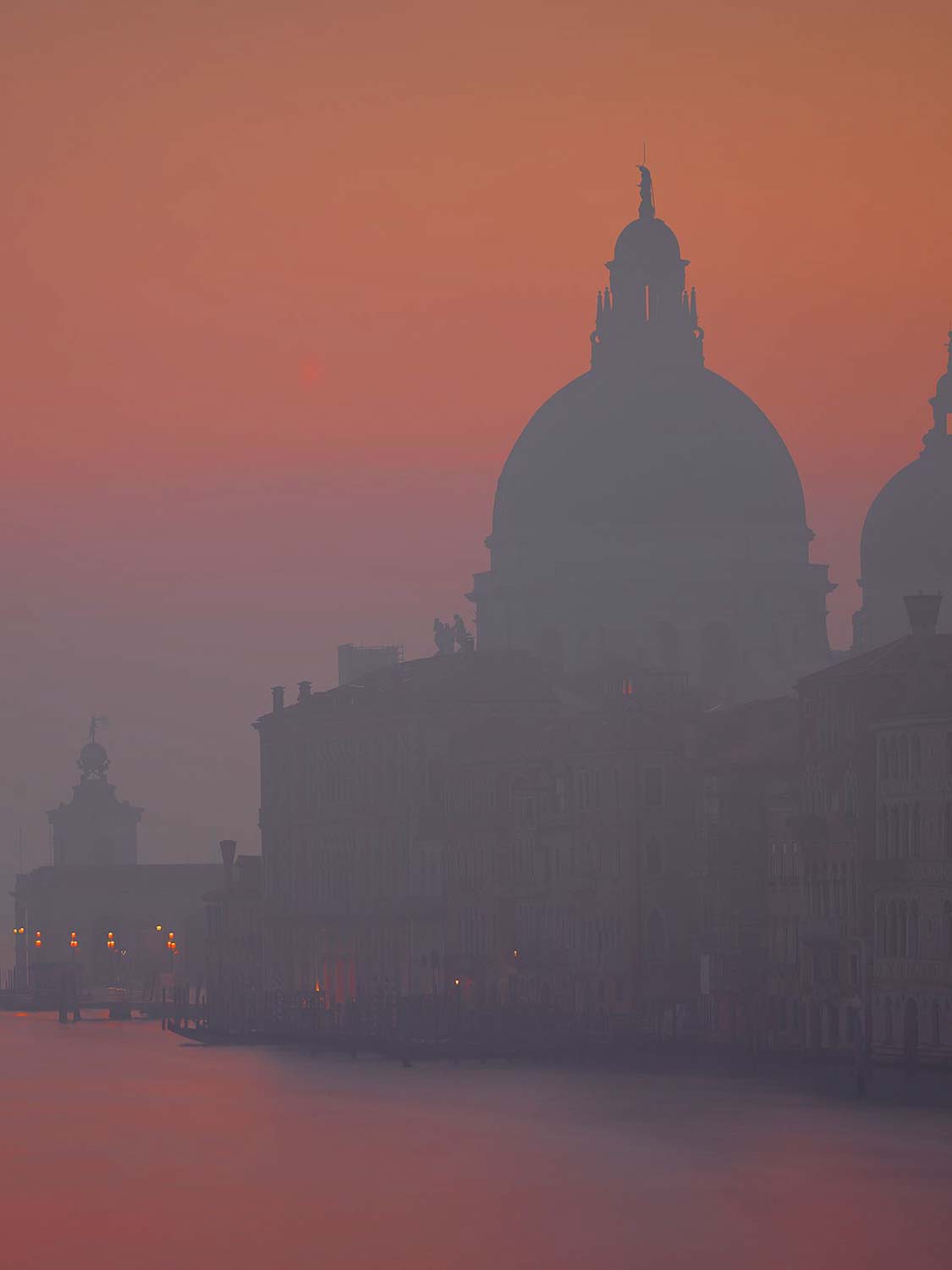 Eine Silhouette der kuppelgekrönten Kirche Santa Maria della Salute in Venedig und der umliegenden Gebäude bei Sonnenuntergang, mit einem sanft orange-rosa gefärbten Himmel und sanften Spiegelungen auf dem Wasser.