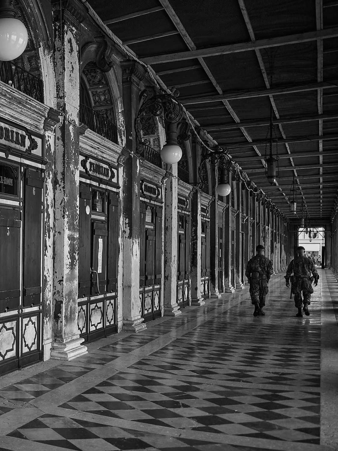 Zwei Menschen in Uniform gehen eine lange, überdachte Arkade mit verzierten Säulen und verwitterten Wänden entlang, deren Schatten auf den karierten Boden fallen; die Szene ist in Schwarz-Weiß gehalten.