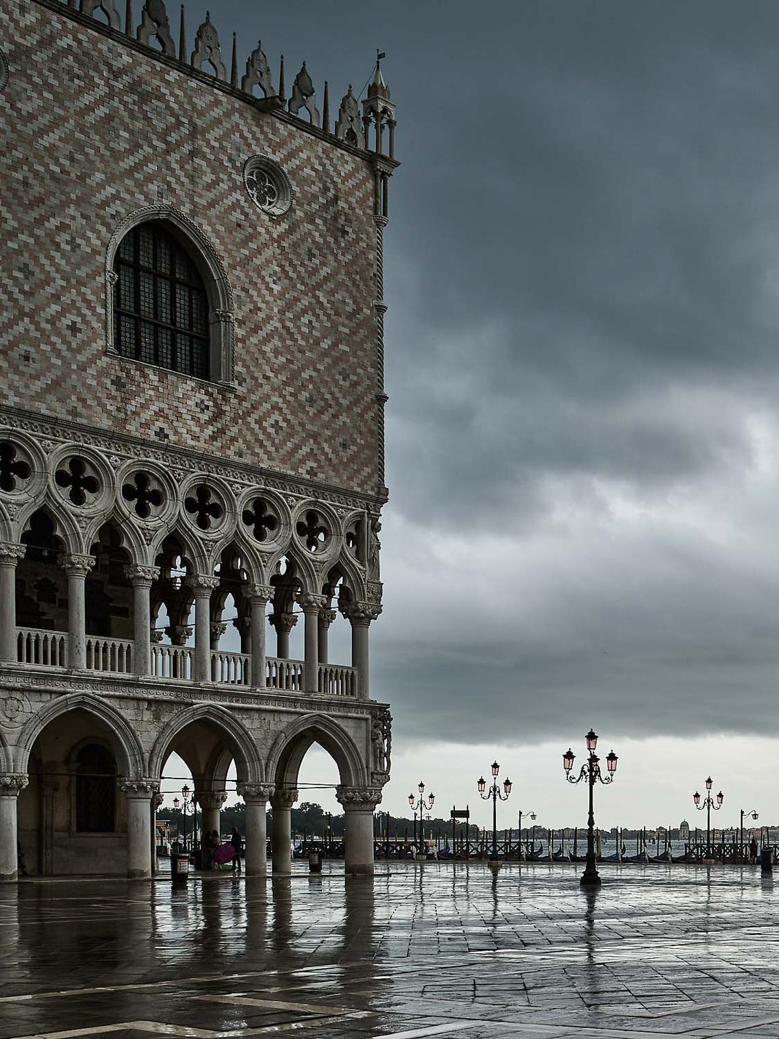 Ein Blick auf den Dogenpalast in Venedig mit seinen kunstvollen Bögen, dem nassen Pflaster, in dem sich die Straßenlaternen spiegeln, und einem bewölkten Himmel über der Uferpromenade im Hintergrund.
