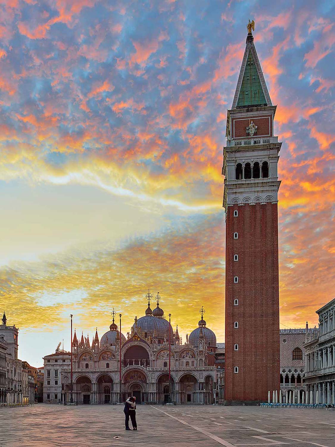 Der Markusplatz in Venedig bei Sonnenaufgang: Der hohe Campanile-Turm und die kunstvoll verzierten Kuppeln der Markusbasilika vor einem farbenprächtigen Himmel, während zwei Menschen auf dem menschenleeren Platz stehen.
