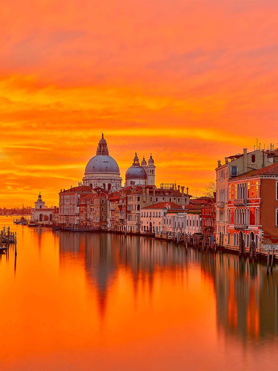 Ein leuchtend orangefarbener Sonnenuntergang über Venedig, Italien, spiegelt sich im Wasser wider. Historische Gebäude und die Kuppelkirche Santa Maria della Salute säumen den Canal Grande und schaffen eine ruhige und malerische Kulisse.