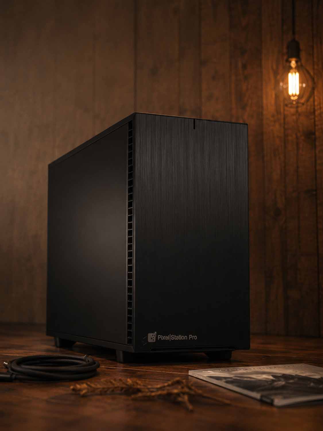 Ein schwarzer PixelStation Pro-Computertower steht auf einer Holzfläche neben einem aufgerollten Kabel und einer Zeitschrift, umgeben von warmem Hängelicht und holzvertäfelten Wänden – perfekt für Fotografie oder jedes KI-Projekt in einem innovativen IT-Systemhaus.