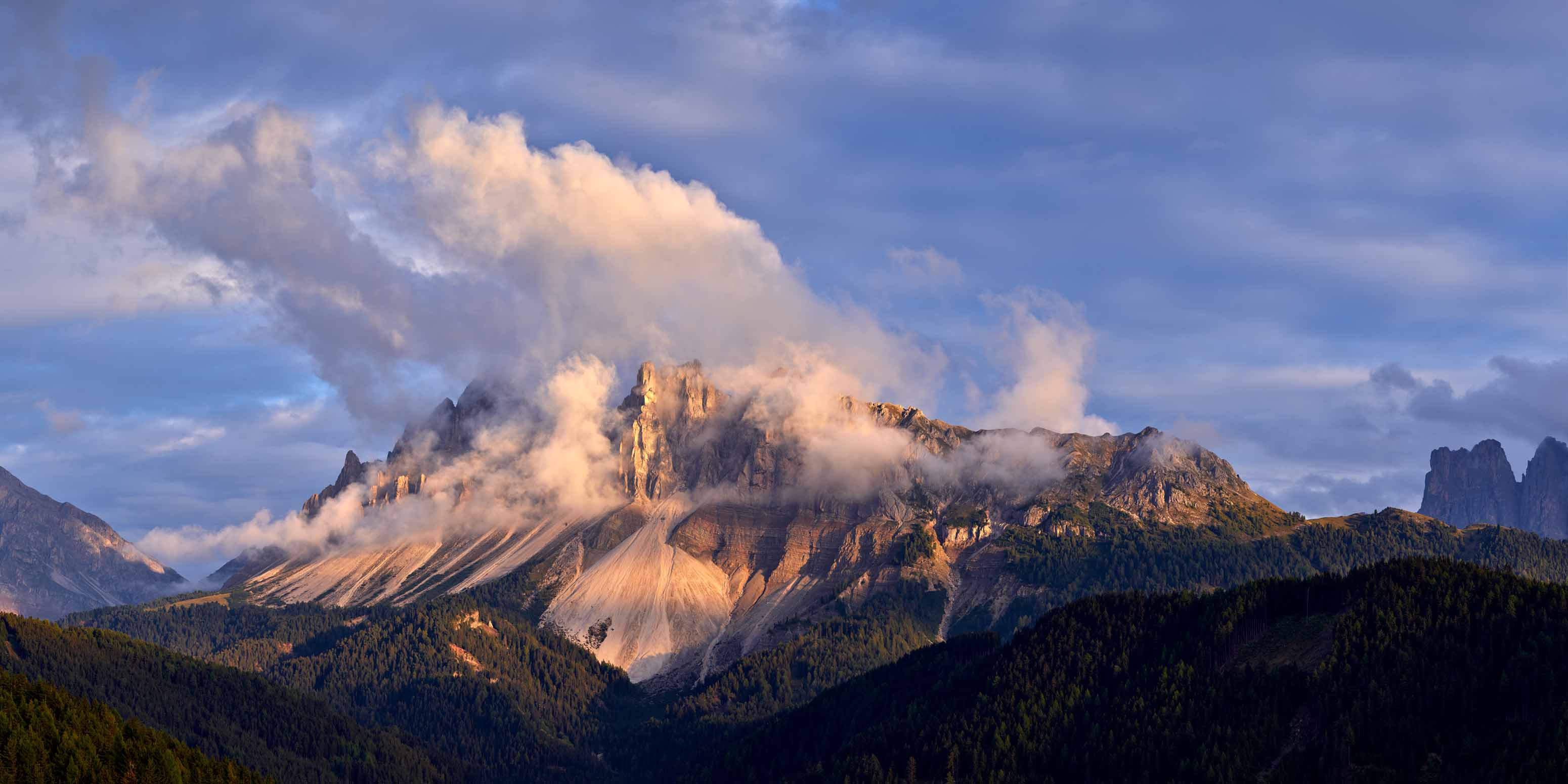 Felsige Berggipfel, die teilweise von dramatischen Wolken verdeckt werden, mit Sonnenlicht, das die Hänge und dichte grüne Wälder im Vordergrund unter einem blauen Himmel beleuchtet - fachmännisch aufgenommen von Phase One Certified Pro Europa Frank Werner Bildbearbeitung.