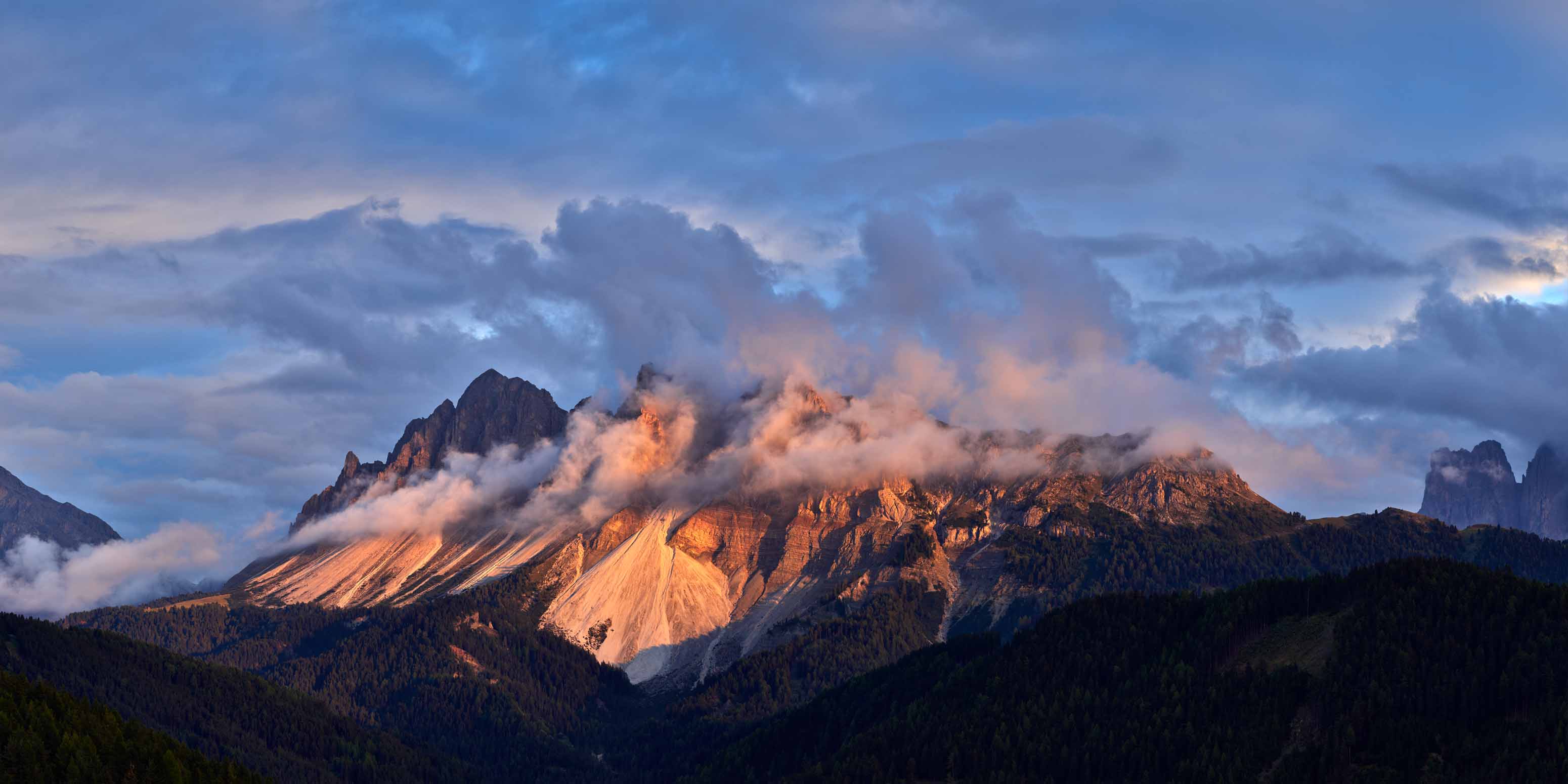 Schneebedeckte Berge, die teilweise von Wolken bedeckt sind, werden in warmes Sonnenlicht getaucht, mit Kiefernwäldern im Vordergrund und einem dramatischen, farbenfrohen Himmel über ihnen - aufgenommen von Frank Werner Fotograf, Phase One Certified Pro Europa.