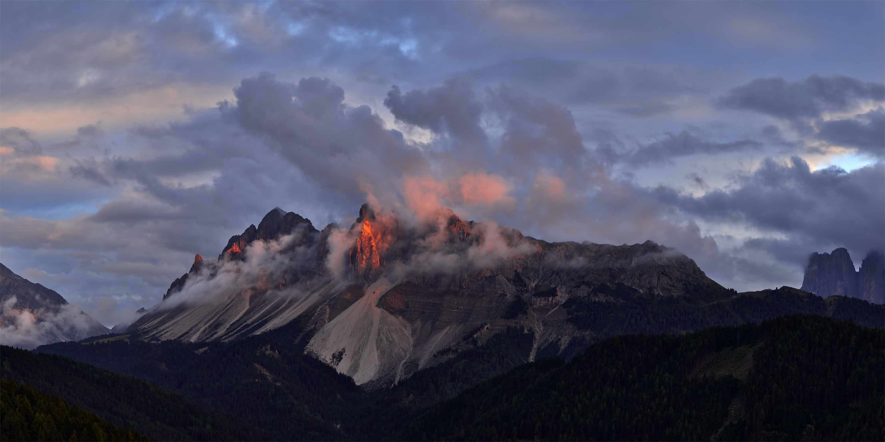 Wolken wirbeln um zerklüftete Berggipfel, während warmes Sonnenlicht die felsigen Gipfel beleuchtet. Die von Frank Werner Fotograf, Phase One Certified Pro Europa, aufgenommene Szene zeigt dichte Wälder und schroffe Hänge unter einem dramatischen, teilweise bewölkten Himmel.